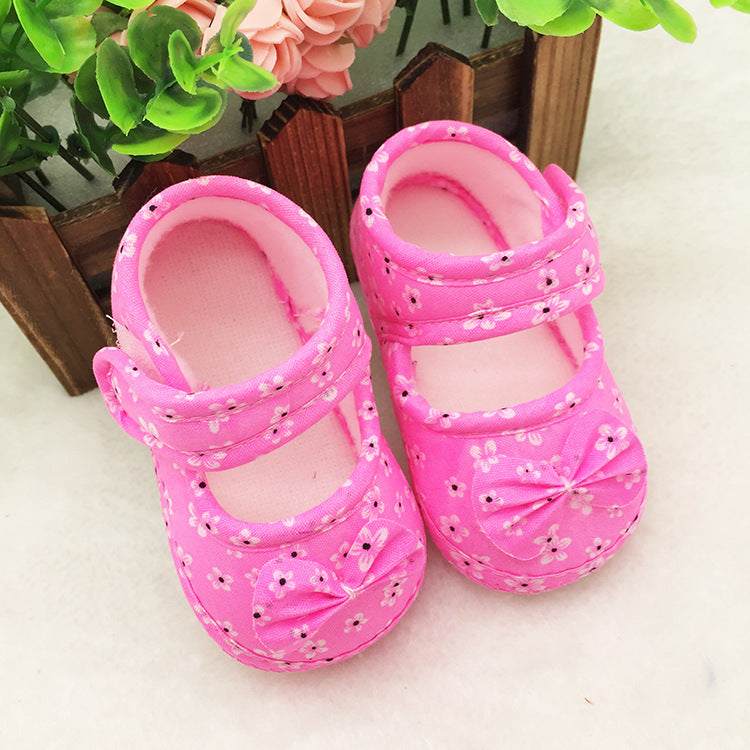 Pink floral baby shoes with bows sit on a white surface. They are placed near a wooden box adorned with green leaves and pink roses.