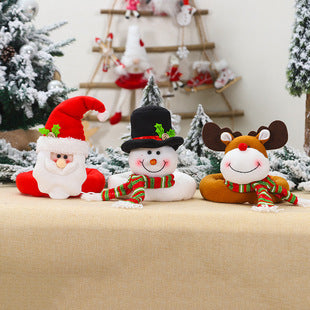 Festive plush toys of Santa, a snowman, and a reindeer with striped scarves sit on a beige surface. Snow-covered trees and holiday decorations are in the background.