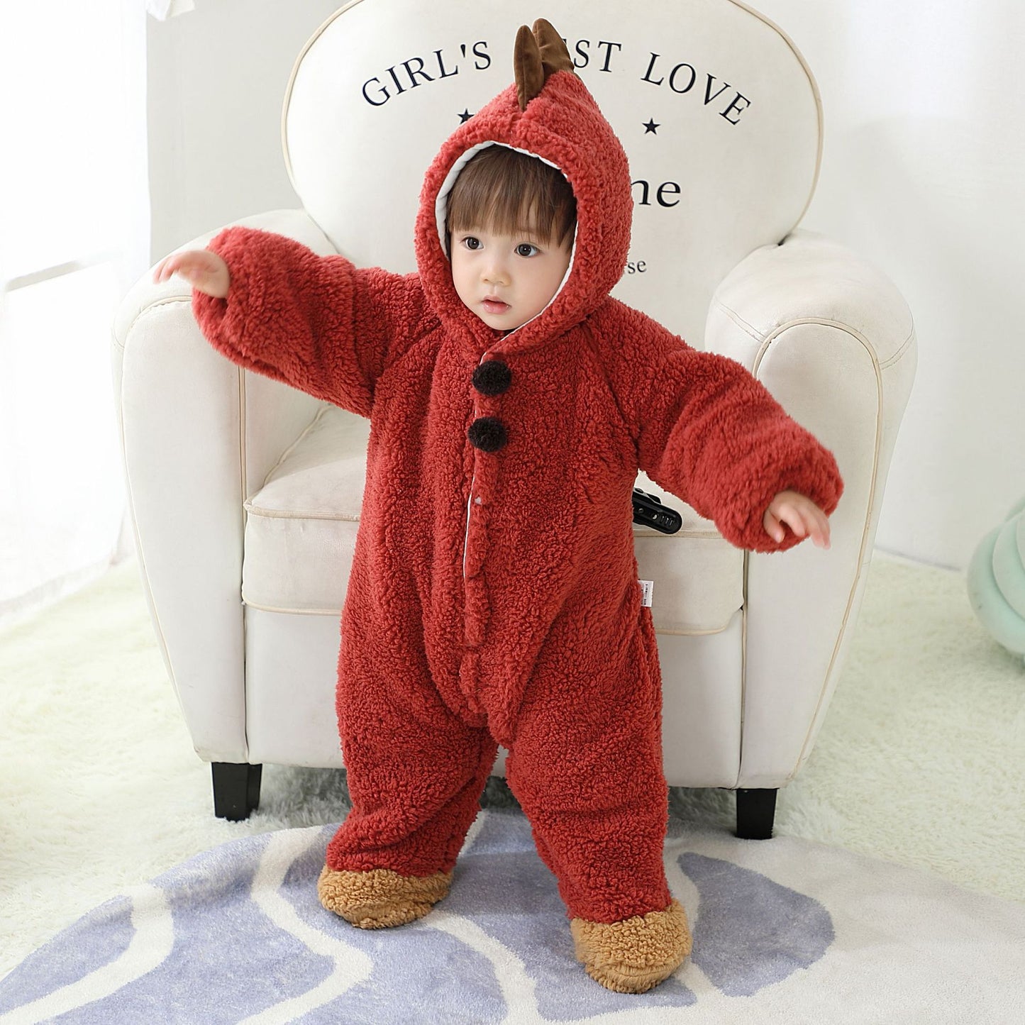 A toddler in a fluffy red onesie with a hood and bear-style ears stands on a rug, arms outstretched. The child is in front of a white armchair with text on it.