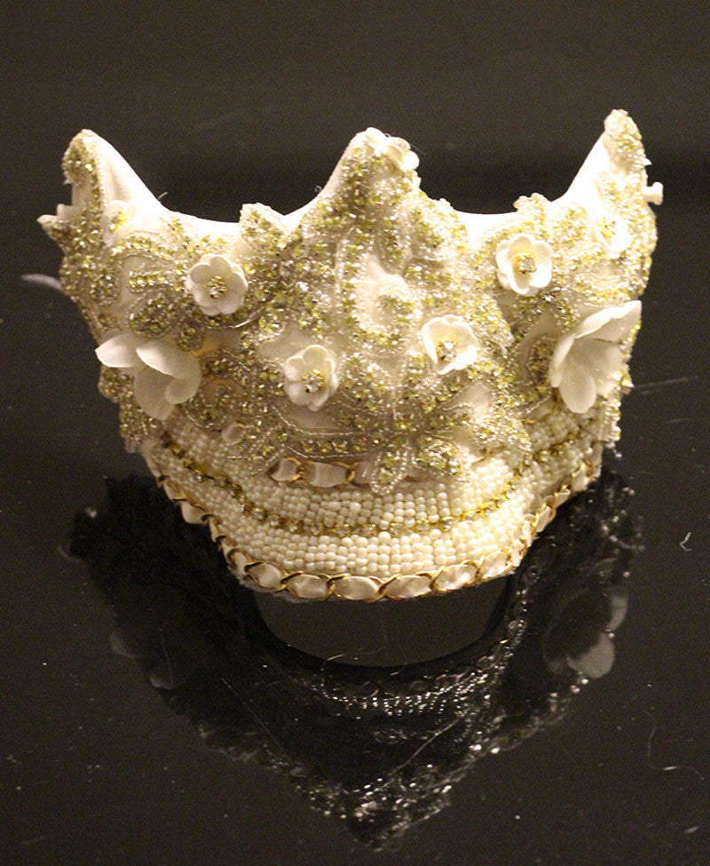 Ornate white and gold decorative mask with intricate beadwork and floral details sits on a glossy black surface, reflecting light.