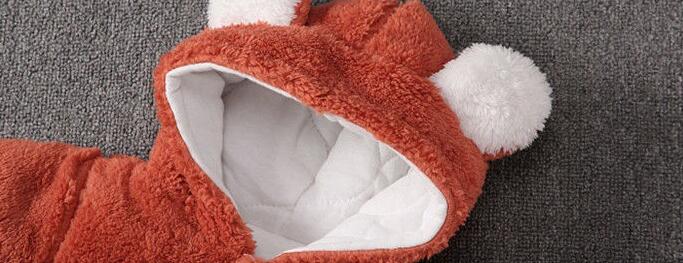 Brown fuzzy hooded garment with white lining, featuring animal-like ears topped with white pom-poms, laid on a textured gray surface.