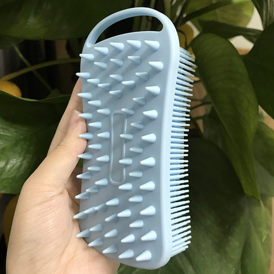A hand holding a light blue silicone brush with a handle. One side features large spikes, while bristles cover the other. Lush green leaves provide the background.