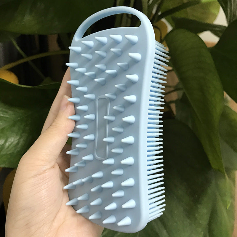 A hand holding a blue silicone brush with pointed bristles on one side and finer bristles on the other, used for cleaning or massaging. Background includes green leaves.
