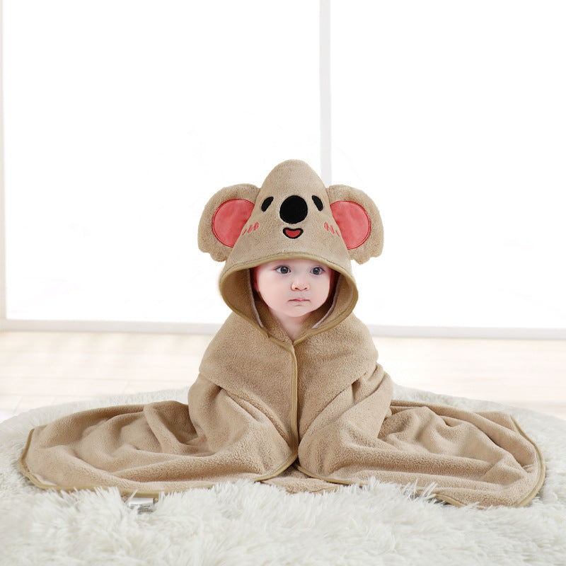A baby wrapped in a brown fleece blanket with a koala design and hood sits on a white fluffy rug. The hood features koala ears and a cheerful face, creating an adorable scene.