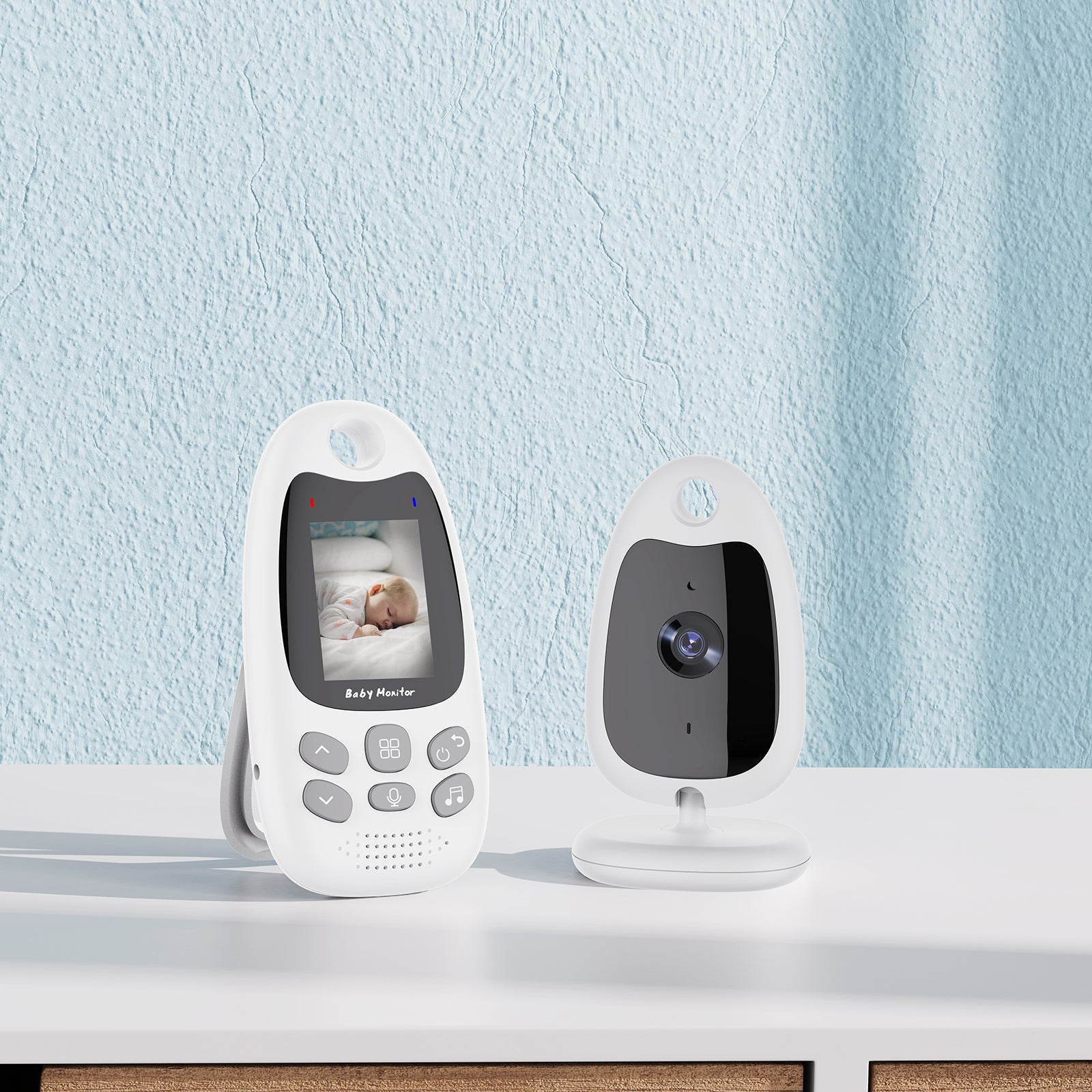 White baby monitor on a table with a camera and a screen showing a sleeping baby. The wall behind is light blue, and the design is sleek and modern.