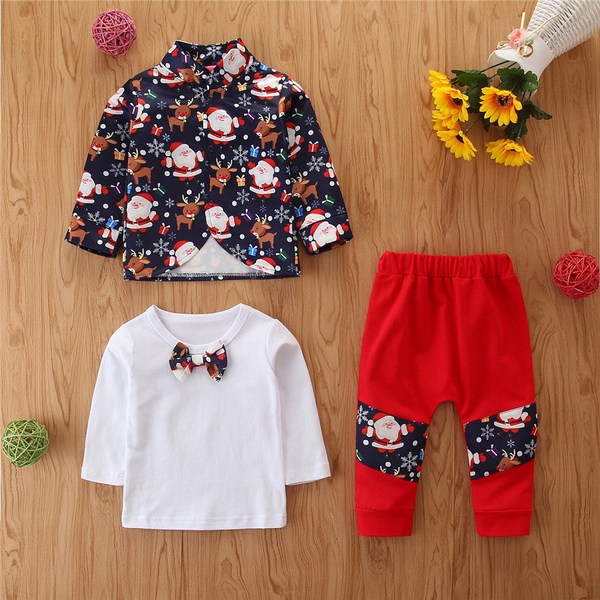 Children's festive outfit set on a wooden surface, featuring a navy jacket with Santa and reindeer, white shirt with bowtie, and red pants with matching festive patches.