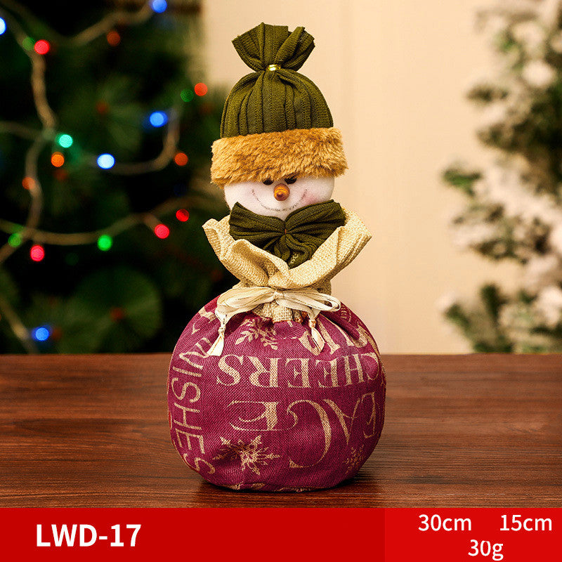 Decorative snowman ornament with a green hat and scarf sits on a table. The purple fabric base has gold lettering. Festive lights and a Christmas tree are blurred in the background.