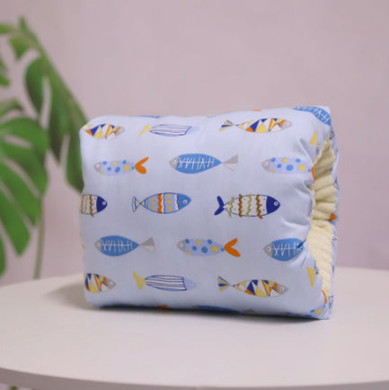 Blue cushion with colorful fish pattern, featuring various shapes and designs, sits on a white surface. A green plant is slightly visible in the background against a pink wall.