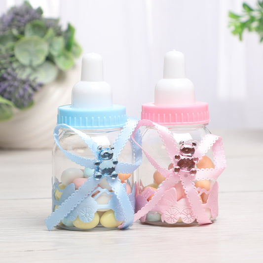 Two baby bottle-shaped containers adorned with blue and pink ribbons, featuring small teddy bears. Each bottle is filled with pastel-colored candies, set on a light background.