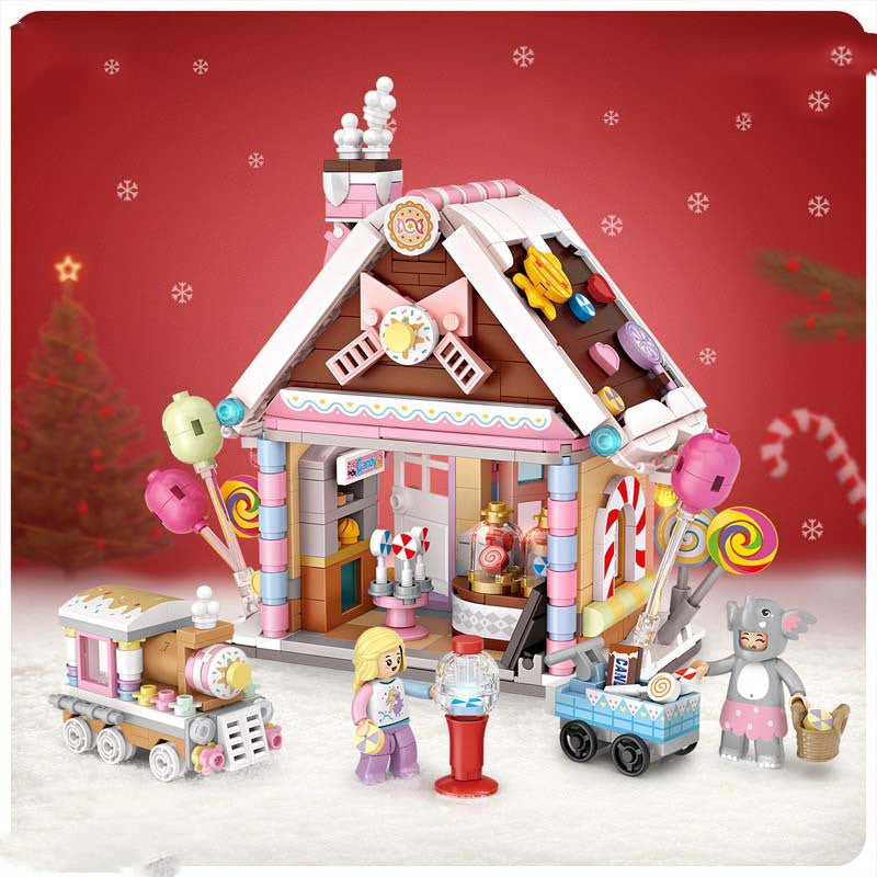 Toy gingerbread house with candy-themed decorations, a mini train, and figurines, set against a snowy red backdrop with snowflakes and a Christmas tree in the distance.