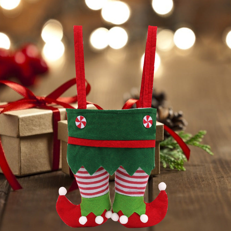 Festive elf-themed gift bag in green with red-and-white striped legs and curly shoes, set on a wooden table with blurred lights and a wrapped present in the background.
