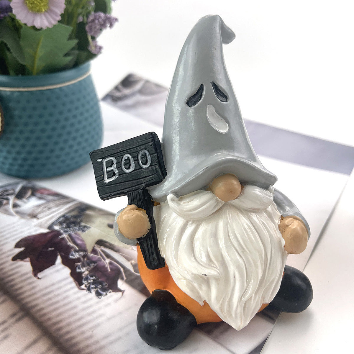 A gnome with a ghost hat, holding a "Boo" sign, is seated on a pumpkin. It is next to a teal pot with flowers on a table with magazine pages.
