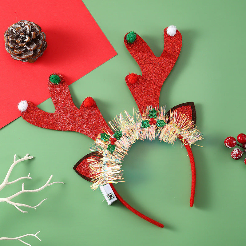 Festive reindeer antler headband with red glitter and pom-poms, decorated with tinsel and holly, on a green and red background with a pinecone and red berries.