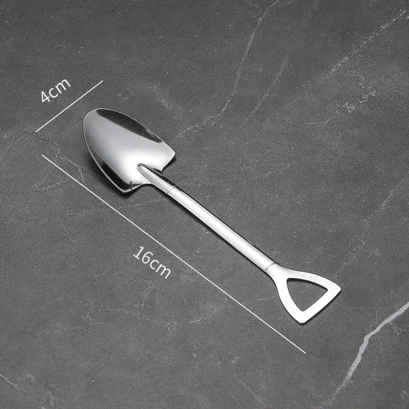 A silver spoon shaped like a shovel with dimensions 4cm wide and 16cm long, placed on a dark, textured surface. The handle is designed like a spade's grip.