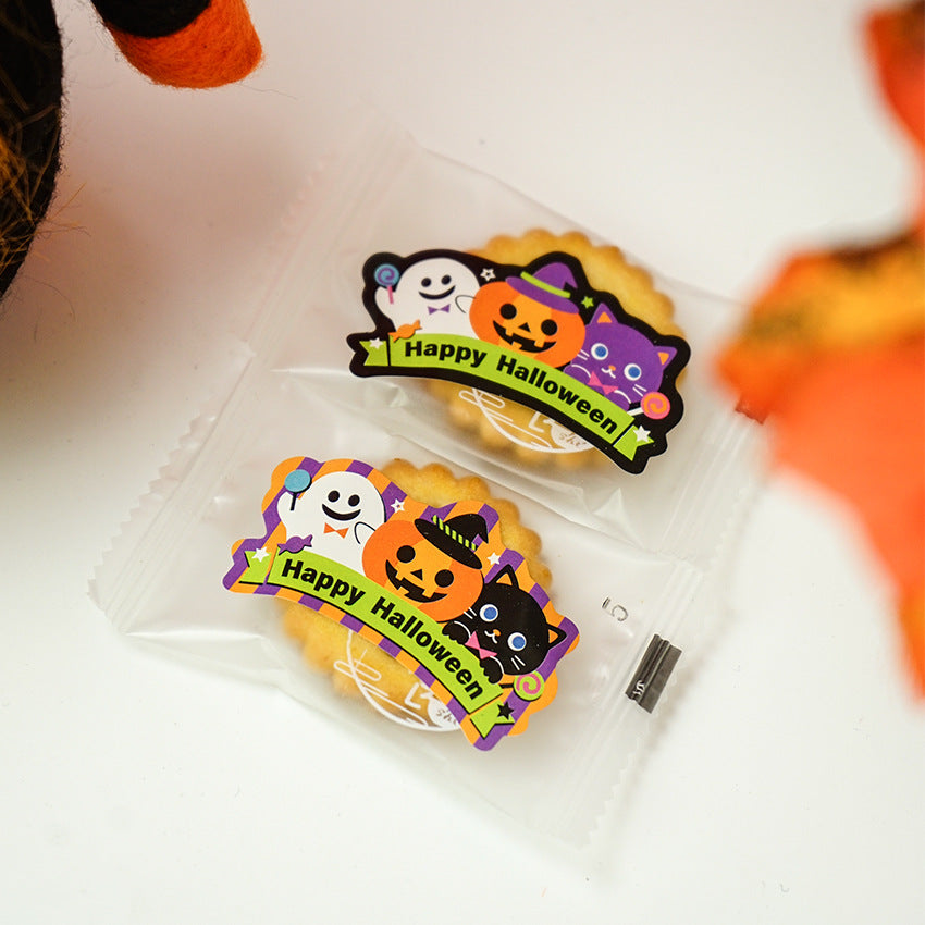 Two Halloween-themed cookies in clear packaging. Each cookie is decorated with a label featuring a cheerful ghost, a pumpkin with a witch hat, and a black cat. The label reads "Happy Halloween" in bold letters. The background has orange and black accents, emphasizing the festive theme.