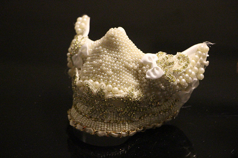Decorative face mask adorned with white pearls, gold glitter, and ribbon bows, set against a dark background.