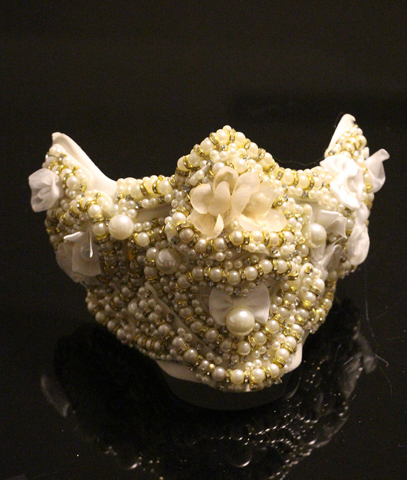 An ornate face mask adorned with white and gold beads, pearls, and fabric flowers against a dark background, creating an elegant, decorative appearance.