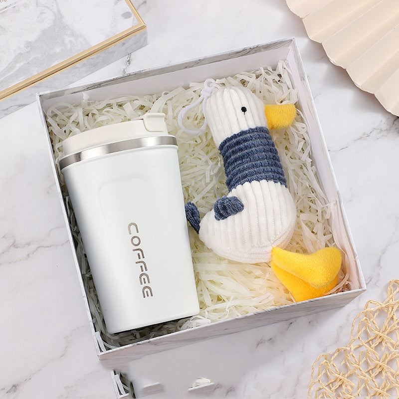 A white travel coffee mug and a knitted duck toy with blue and yellow accents are nestled in crinkled paper inside a decorative box on a marble surface.