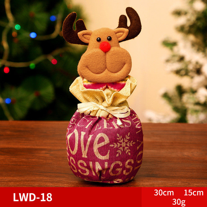 Festive reindeer decoration with a plush face and antlers, red nose, atop a fabric bag with "Love Blessings" text. It sits on a wooden surface with Christmas lights in the background.