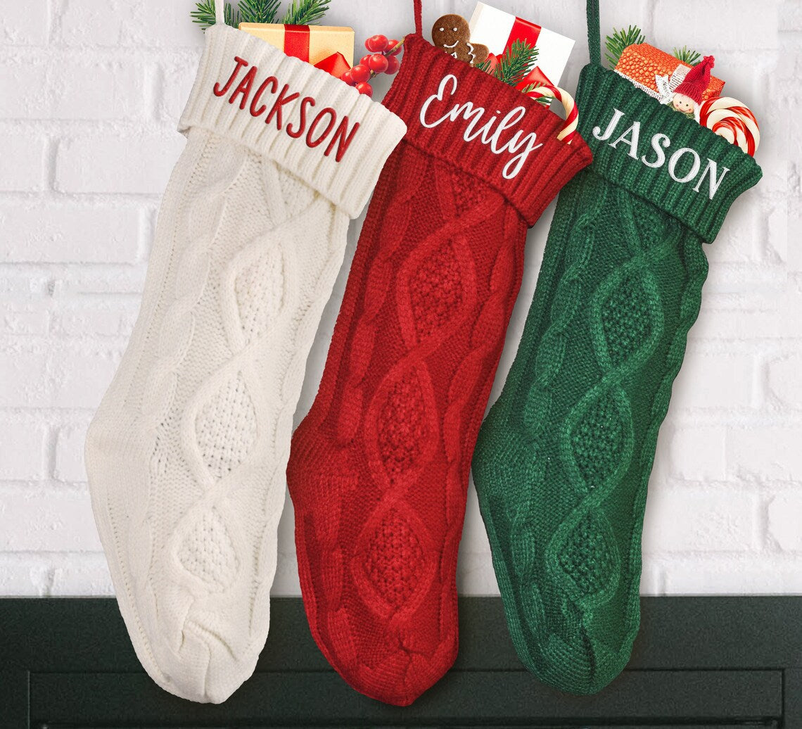 Three knitted Christmas stockings hang against a white brick wall. They are labeled "Jackson" in white, "Emily" in red, and "Jason" in green. They are filled with gifts and candy.