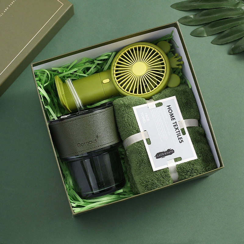 Gift box containing a green handheld fan, a green insulated tumbler labeled "Gorgeous," and a rolled green towel with a "Home Textiles" tag, on green shredded paper.