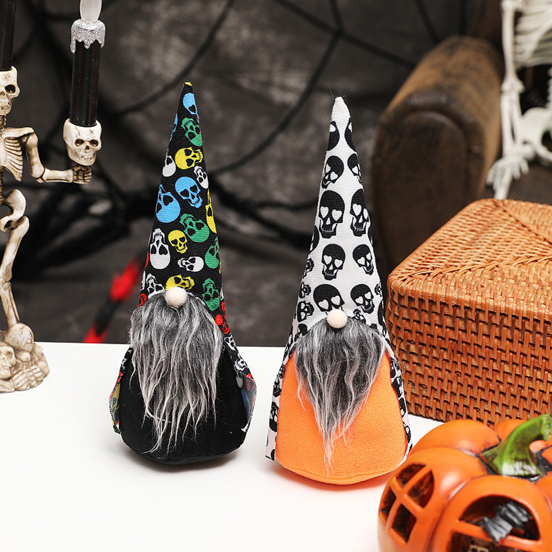 Two Halloween gnomes with long gray beards and pointy hats featuring skull patterns sit on a table. One wears a multicolored hat and black body, the other has a black-and-white hat and orange body. A wicker basket, skeleton candle holder, and decorative pumpkin are nearby, with a cobweb backdrop.