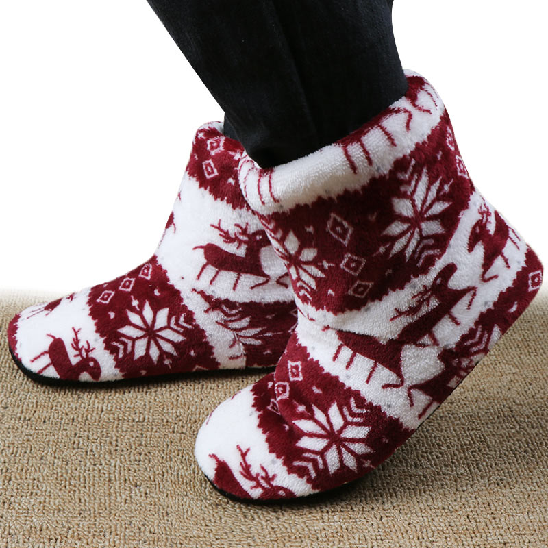 Red and white fuzzy slipper boots with reindeer and snowflake patterns, worn on feet, standing on a textured beige carpet.