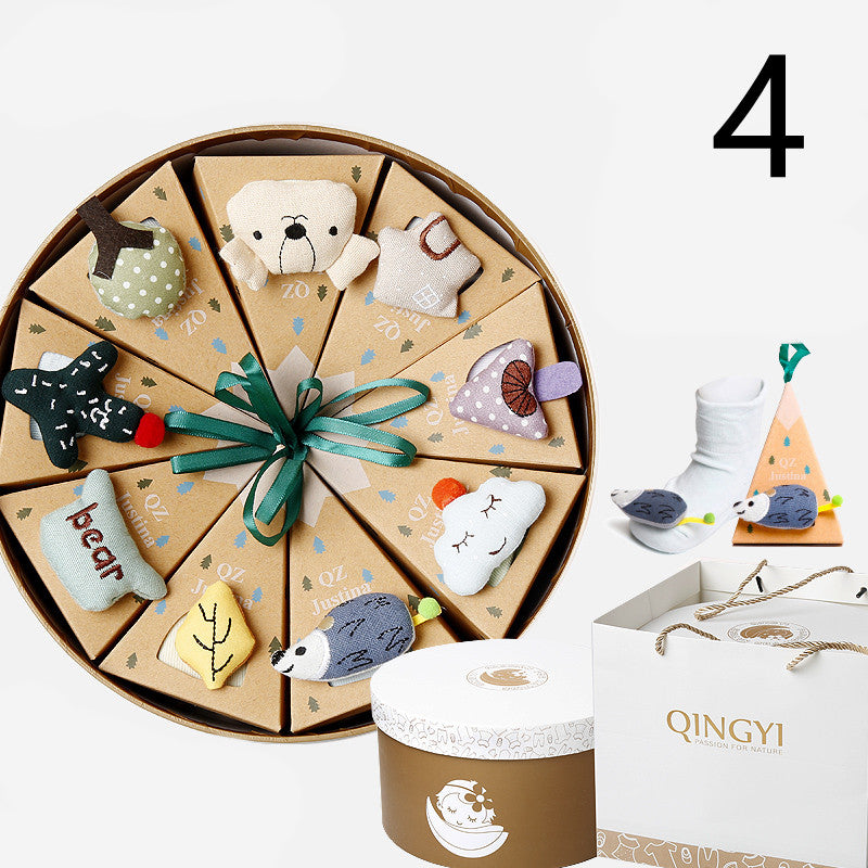 Round gift box with pie-shaped segments, each topped with a soft toy including animals and shapes. Nearby, a sock and animal toy beside a pyramid box. Branded packaging in the foreground.