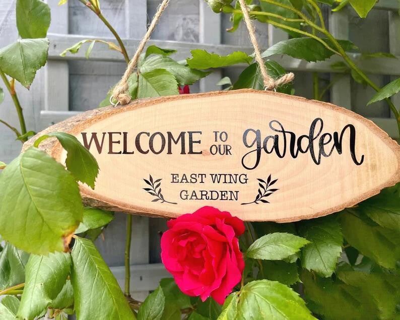 A wooden sign reading "Welcome to our garden, East Wing Garden" hangs among green leaves and a vibrant red rose.