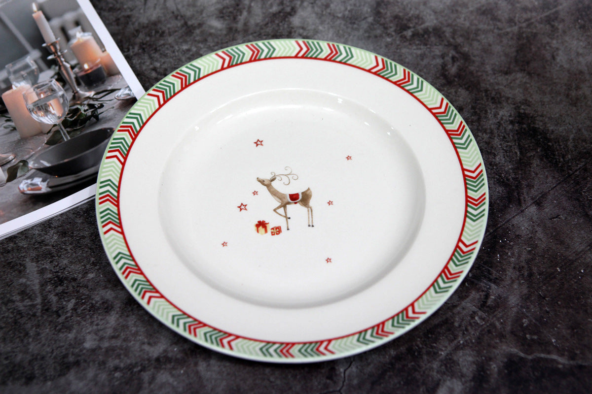 Festive plate with a reindeer and gift illustration in the center, surrounded by red stars. The rim has a red and green chevron pattern. A candlelit table setting is partially visible beside it.
