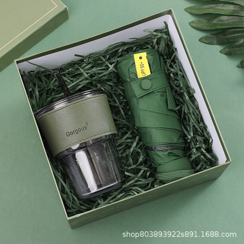 Green gift box with a reusable tumbler and a compact, foldable umbrella nestled in green shredded paper. The tumbler has a light green sleeve, while the umbrella is dark green.