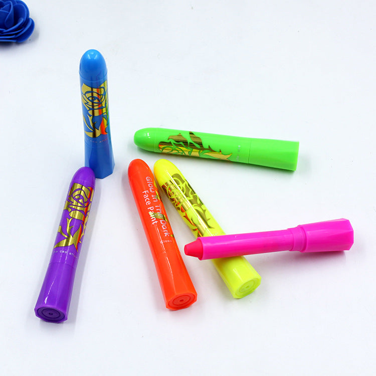Colorful face paint sticks in bright blue, green, purple, orange, yellow, and pink are scattered on a white surface. The orange stick is labeled "glow in the dark."