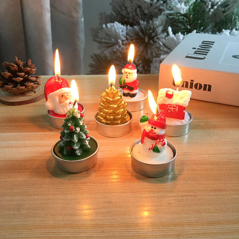 Festive candles shaped like Santa, a snowman, a house, a Christmas tree, and a gold pine cone are lit on a wooden table, with a pine cone and snowy decor in the background.