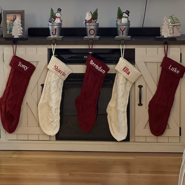 Five Christmas stockings labeled with names hang from a white cabinet. Above, small pots with snowmen and a Santa figure spell "JOY."