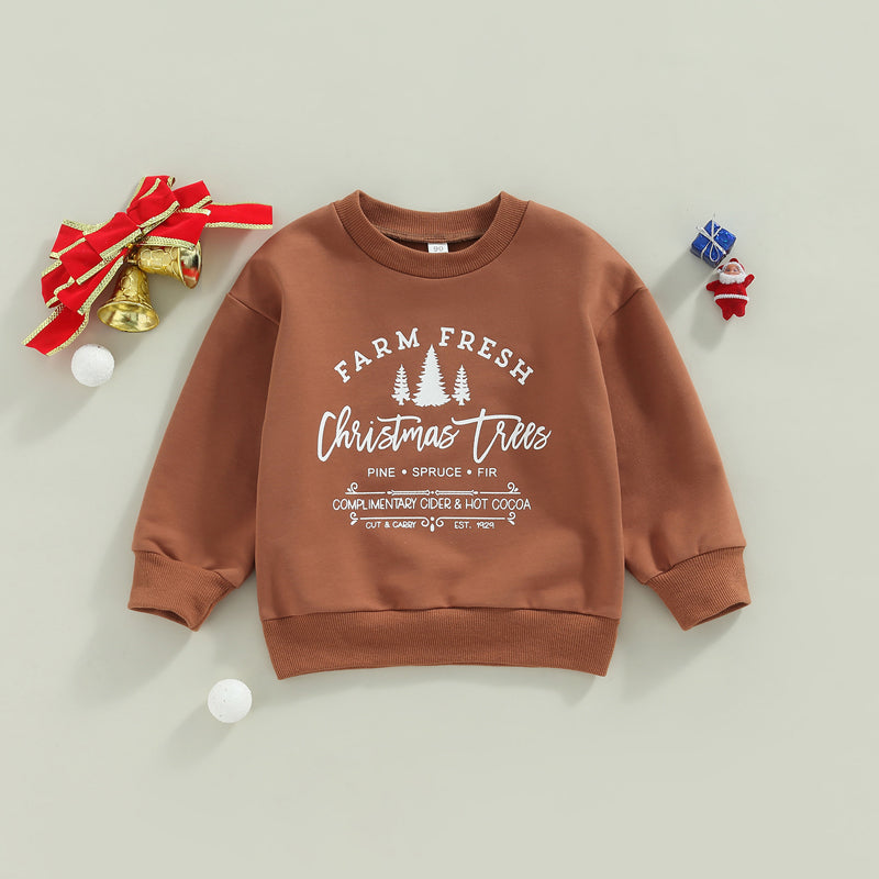 Brown sweatshirt with “Farm Fresh Christmas Trees” print, surrounded by festive decorations: a small Santa figurine, a wrapped gift, golden bells, and white pom-poms on a light background.