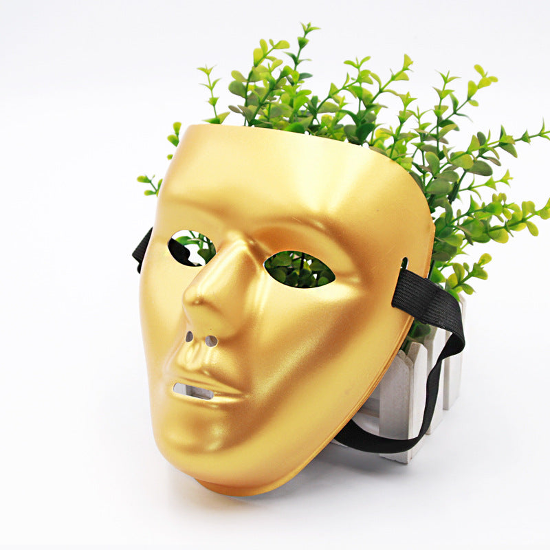 A golden mask lies in front of a pot filled with lush green plants, creating the illusion of hair. The mask is shiny with hollow eyes and a black strap.