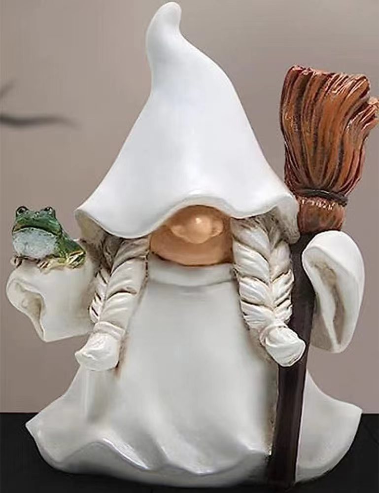 A whimsical ceramic figurine of a gnome with a pointed hat, holding a broom and a frog. The gnome's face is hidden, but has long braided hair, wearing a white robe.