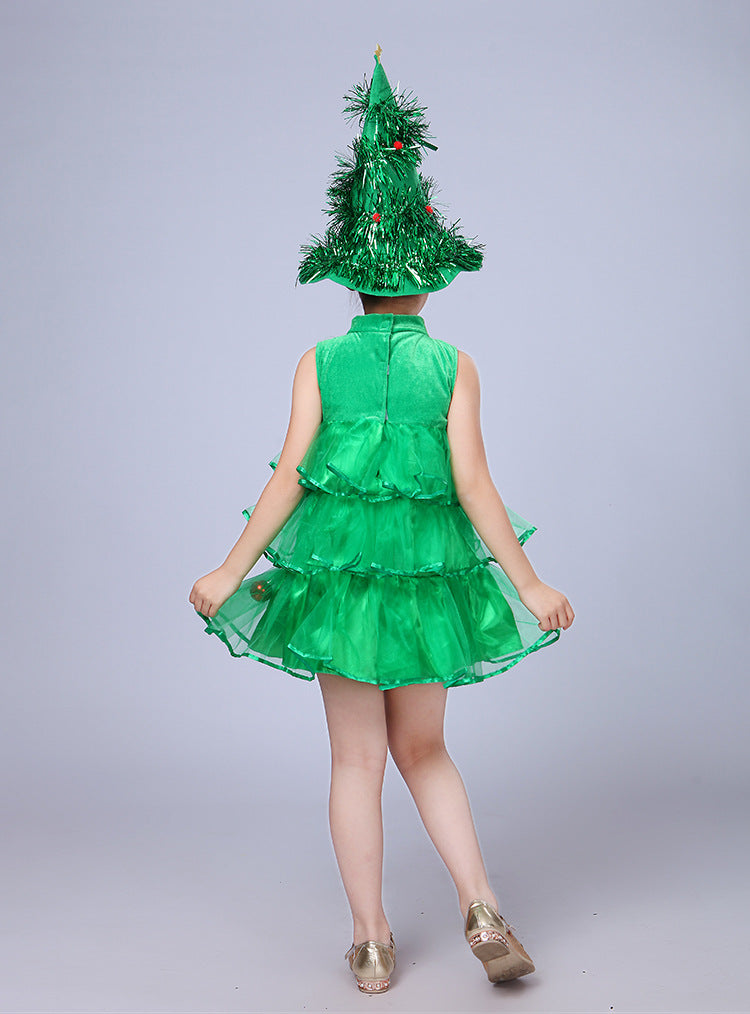 Young child in a layered green dress and a tall, festive Christmas tree hat with red ornaments, facing away. The outfit is playful, evoking a holiday spirit.
