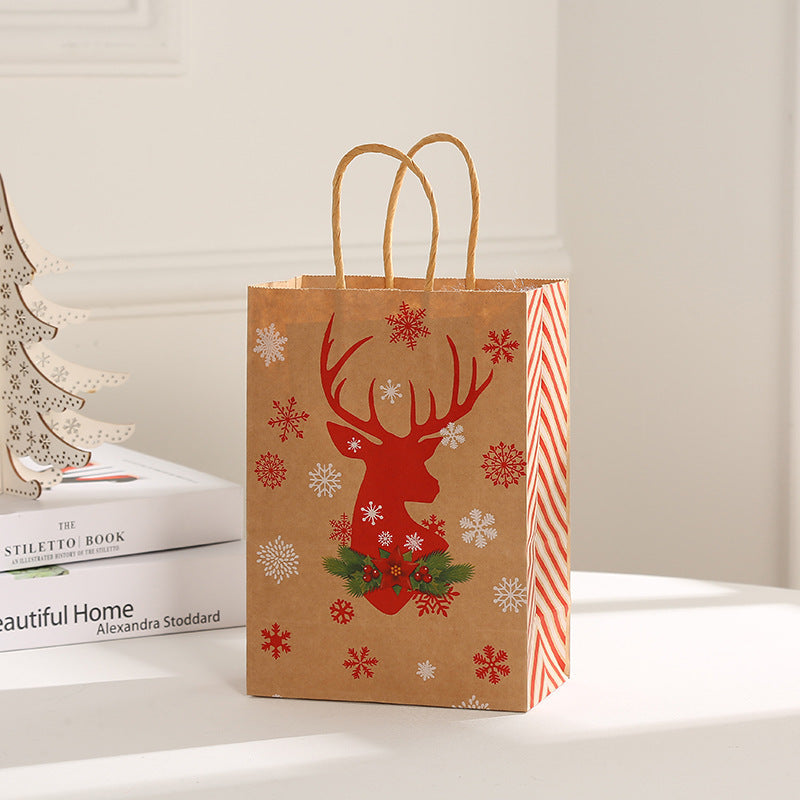 Brown gift bag with a red reindeer silhouette and snowflakes, adorned with green and red holly. It has rope handles and red striped sides, sitting on a white table.