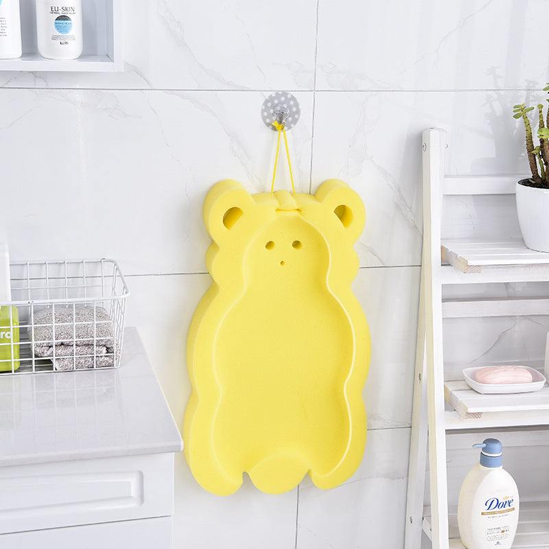 A yellow teddy bear-shaped bath sponge hangs on a tiled bathroom wall. Nearby, shelves hold toiletries, a potted plant, and soap accessories.