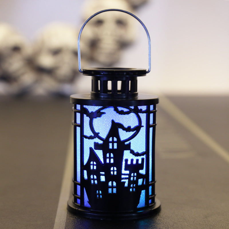 A small, decorative lantern with a black silhouette of a haunted house against an illuminated blue background. The lantern's design features a full moon and flying bats, creating a spooky atmosphere. A wire handle is attached on top. Blurred skull shapes are visible in the background.
