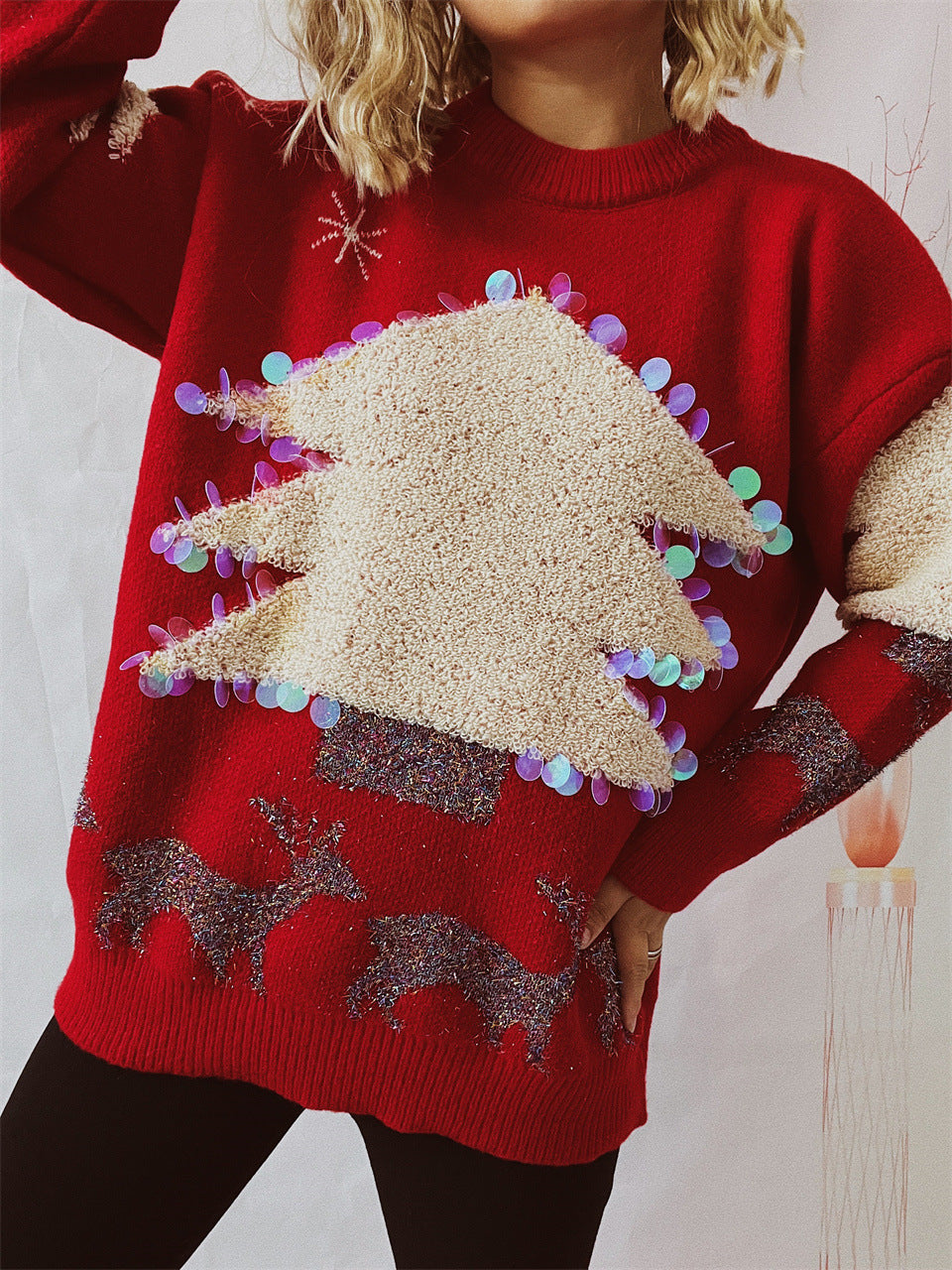 Person wearing a red festive sweater adorned with a large cream Christmas tree and colorful sequins; glittering reindeer patterns decorate the lower part of the sweater.