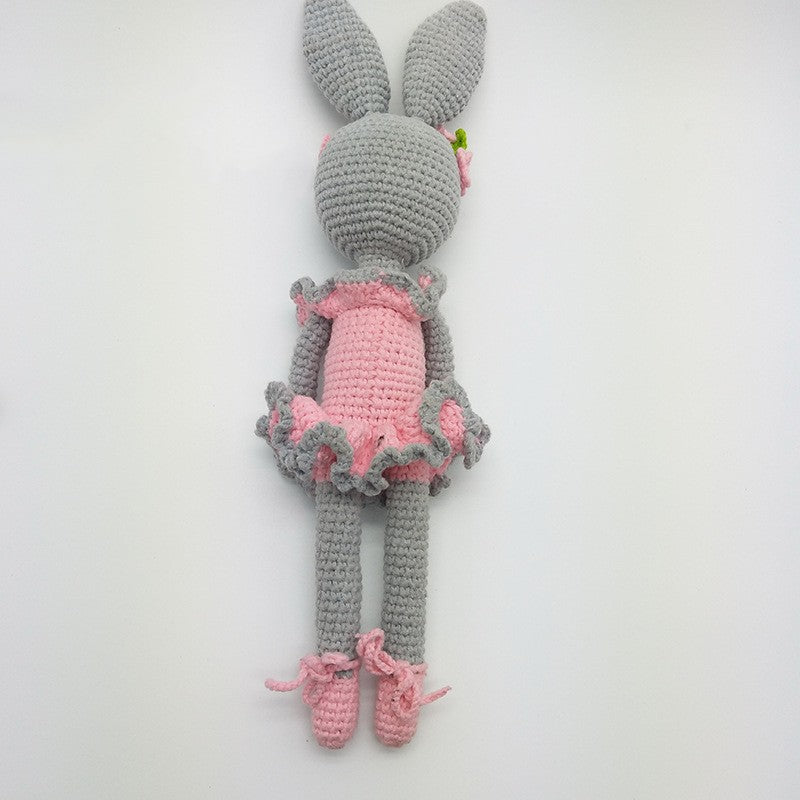 Crocheted gray rabbit doll in a pink ruffled dress and shoes, adorned with a small pink flower. The doll has long ears and is displayed on a neutral background.