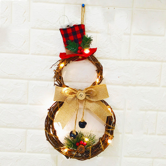 A decorative snowman wreath made from intertwined twigs, with a plaid top hat, burlap bow, pinecones, berries, and lights, hangs on a white brick wall.