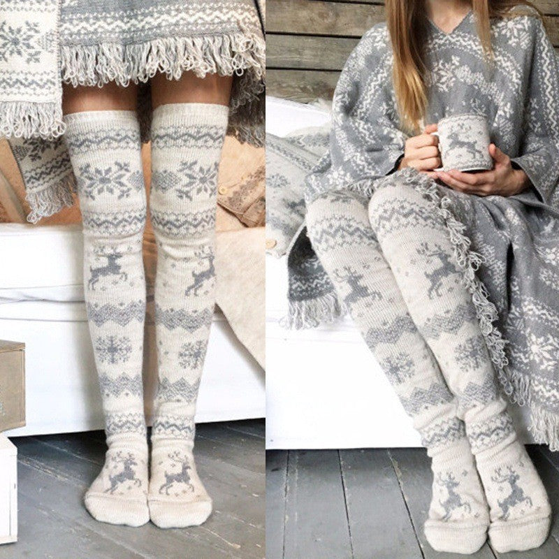 Person wearing cozy, knee-high gray socks and matching fringed poncho with snowflake and reindeer patterns, holding a knitted mug in a rustic room setting.