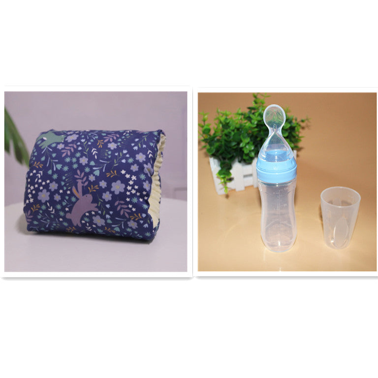 A blue floral-patterned baby pillow and a baby feeding bottle with a spoon-like attachment placed next to a transparent cup on a beige surface with a small plant behind.