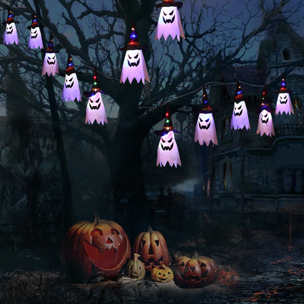 String lights of ghostly figures with colorful hats hang from a tree. Below, carved pumpkins sit on the ground. A spooky, dark house looms in the background.