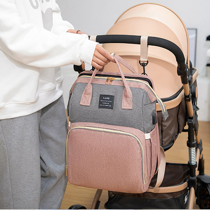 Person in a white sweatshirt and gray pants holding a pink and gray backpack on a stroller handle. The stroller is beige with a black handle in a cozy indoor setting.