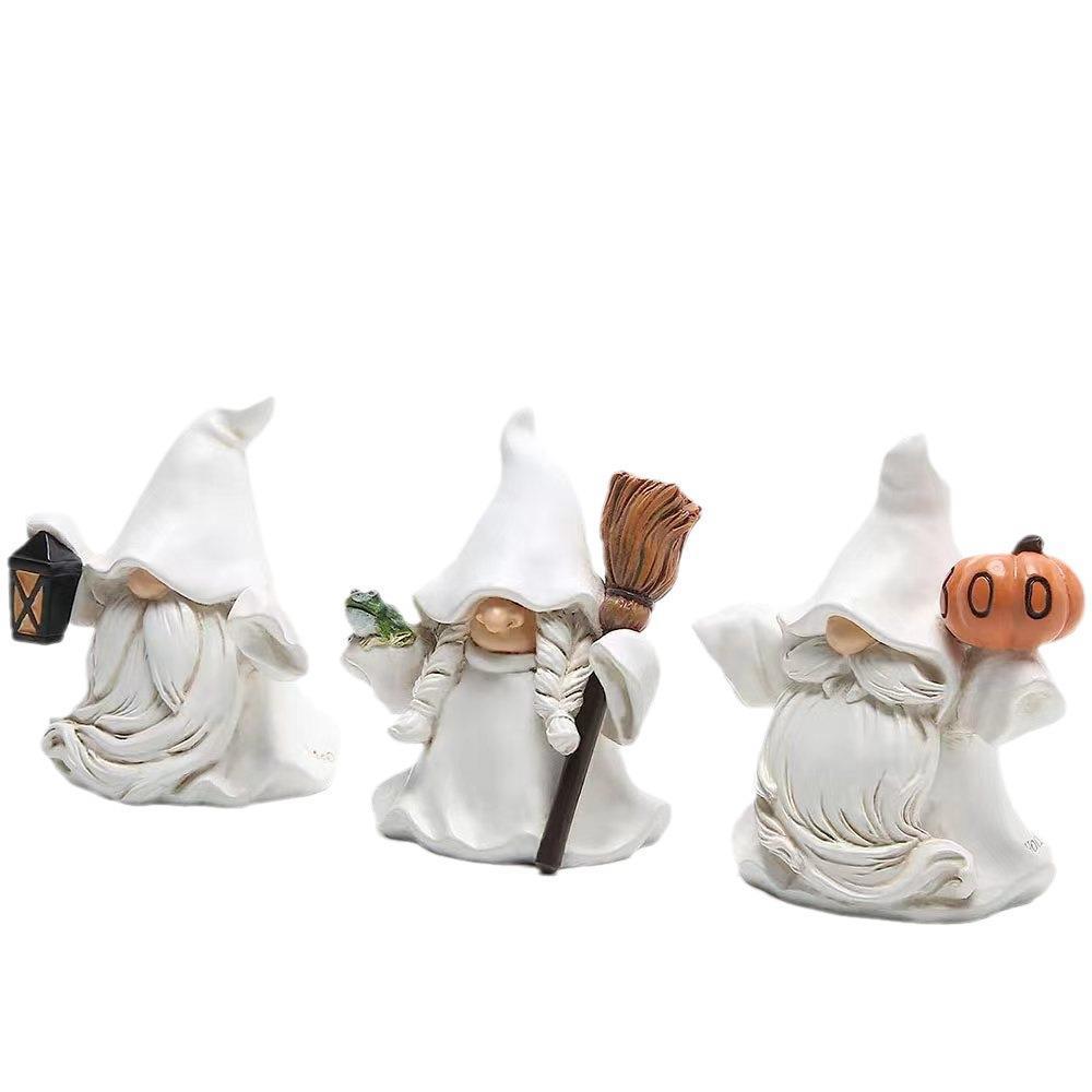 Three small gnome figurines wearing white robes and hats. One holds a lantern, another has a broom and frog, and the third carries a pumpkin.