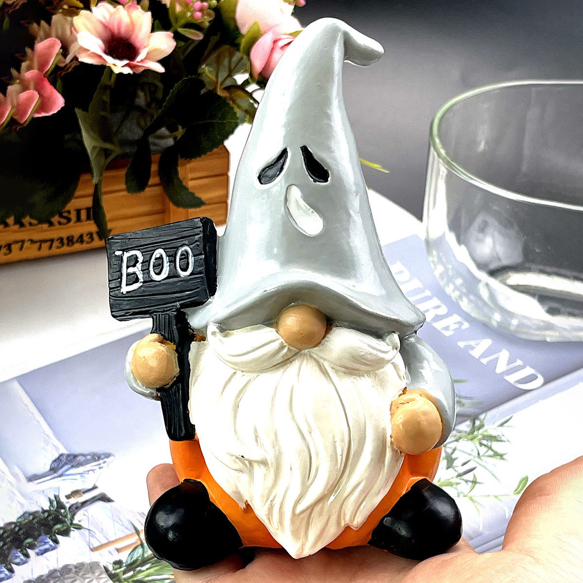 Cute gnome figurine with a ghost-like hat, holding a "Boo" sign. The gnome has a long white beard and sits on an orange base. Flowers in the background.